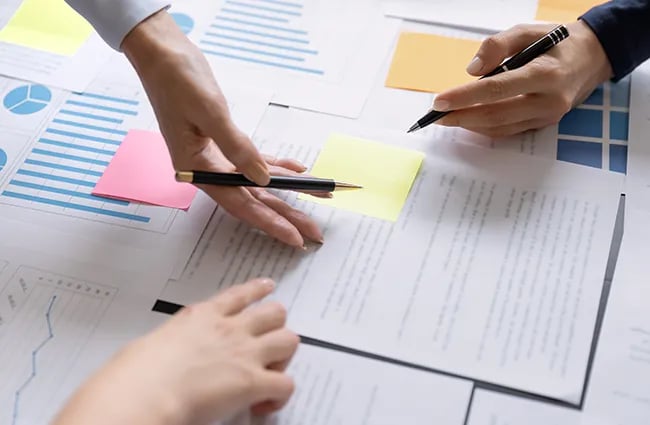Hands holding pens working over layers of papers and post it notes